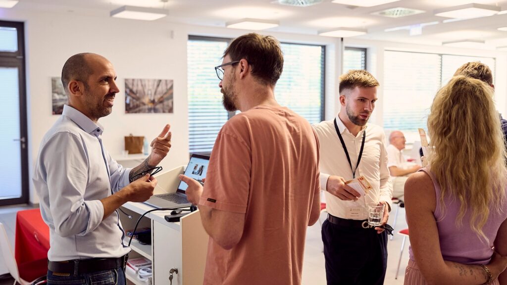 Gruppenszene: vier Erwachsene im Gespräch in einem hellen Büro-/Konferenzraum, einige Personen im Hintergrund.