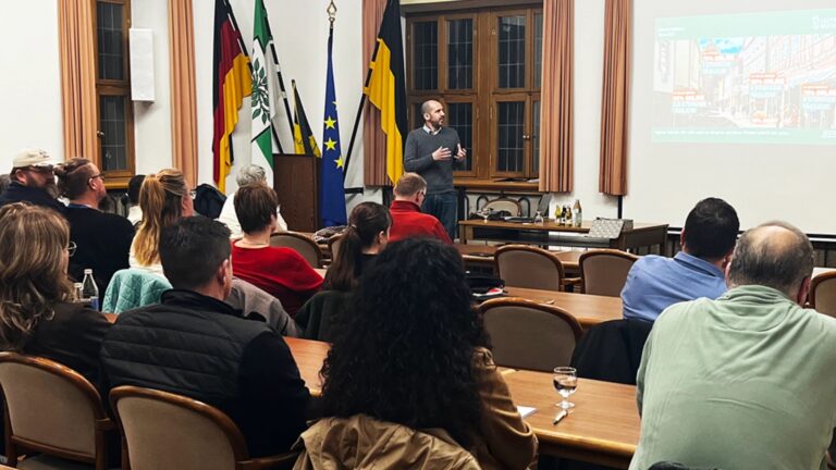 Personen sitzen in einem Konferenzraum und hören einem Referenten vor einer Projektionswand zu, im Hintergrund mehrere Fahnen.